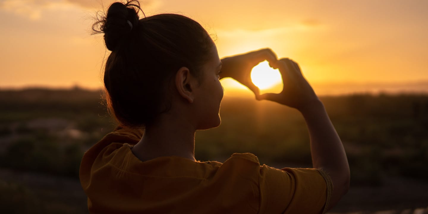 Grateful woman with hands making heart shape in sunset.