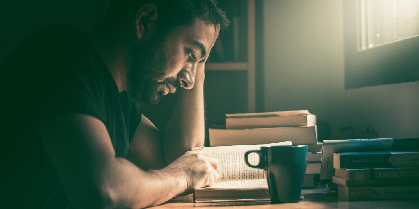 Man sitting at desk reading a book and drinking coffee.