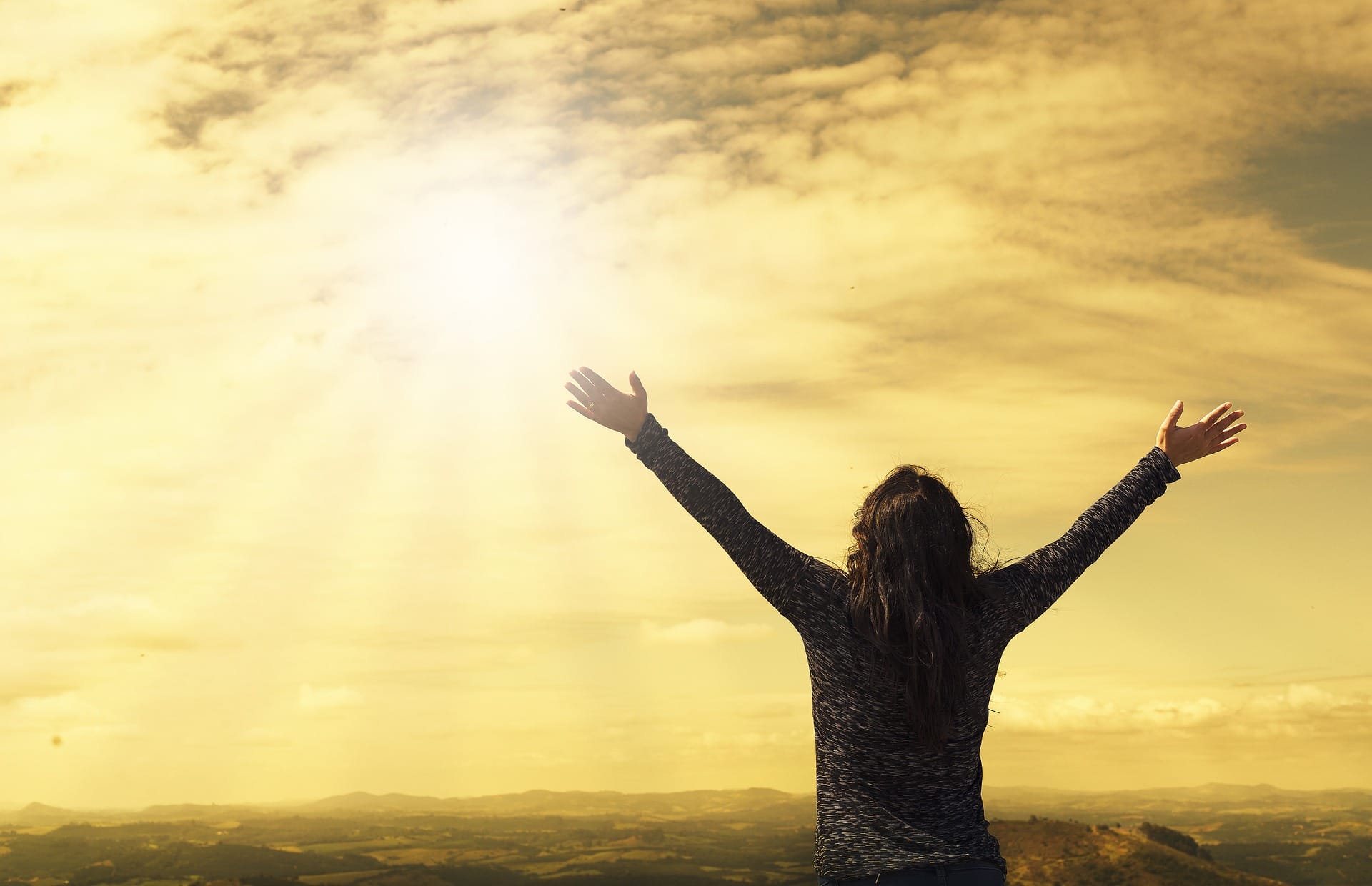 Person holding their arms up in the air and looking towards a sun filled sky.