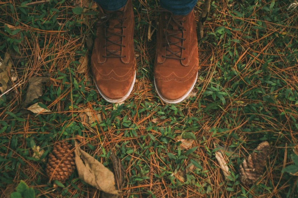 Person's feet wearing brown suede shoes standing on grass and leaves.