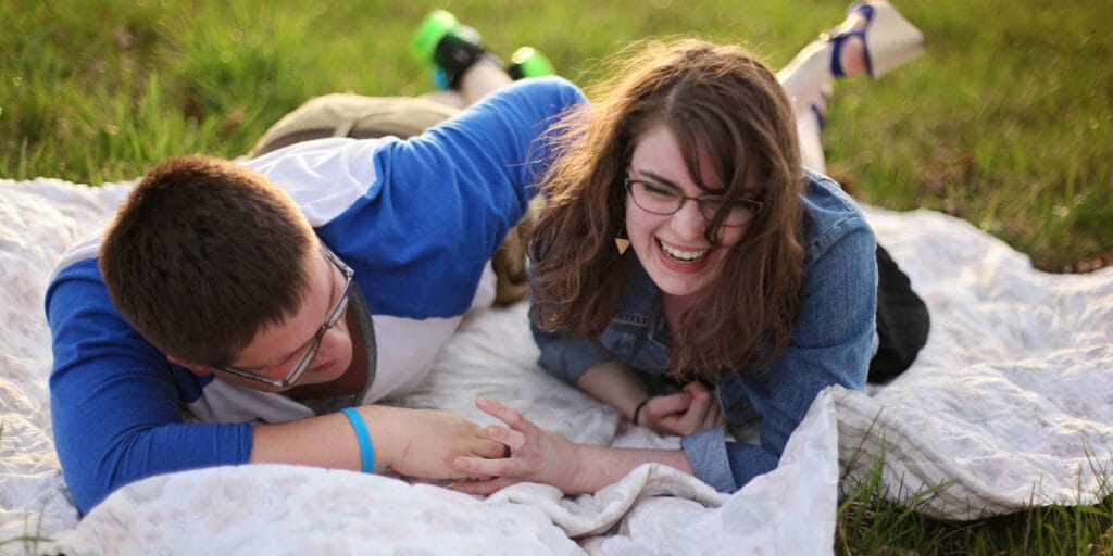 Couple laying on blanket in the grass holding hands and laughing.