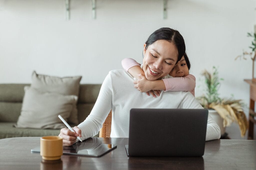 Daughter hugging mother while she is working on computer